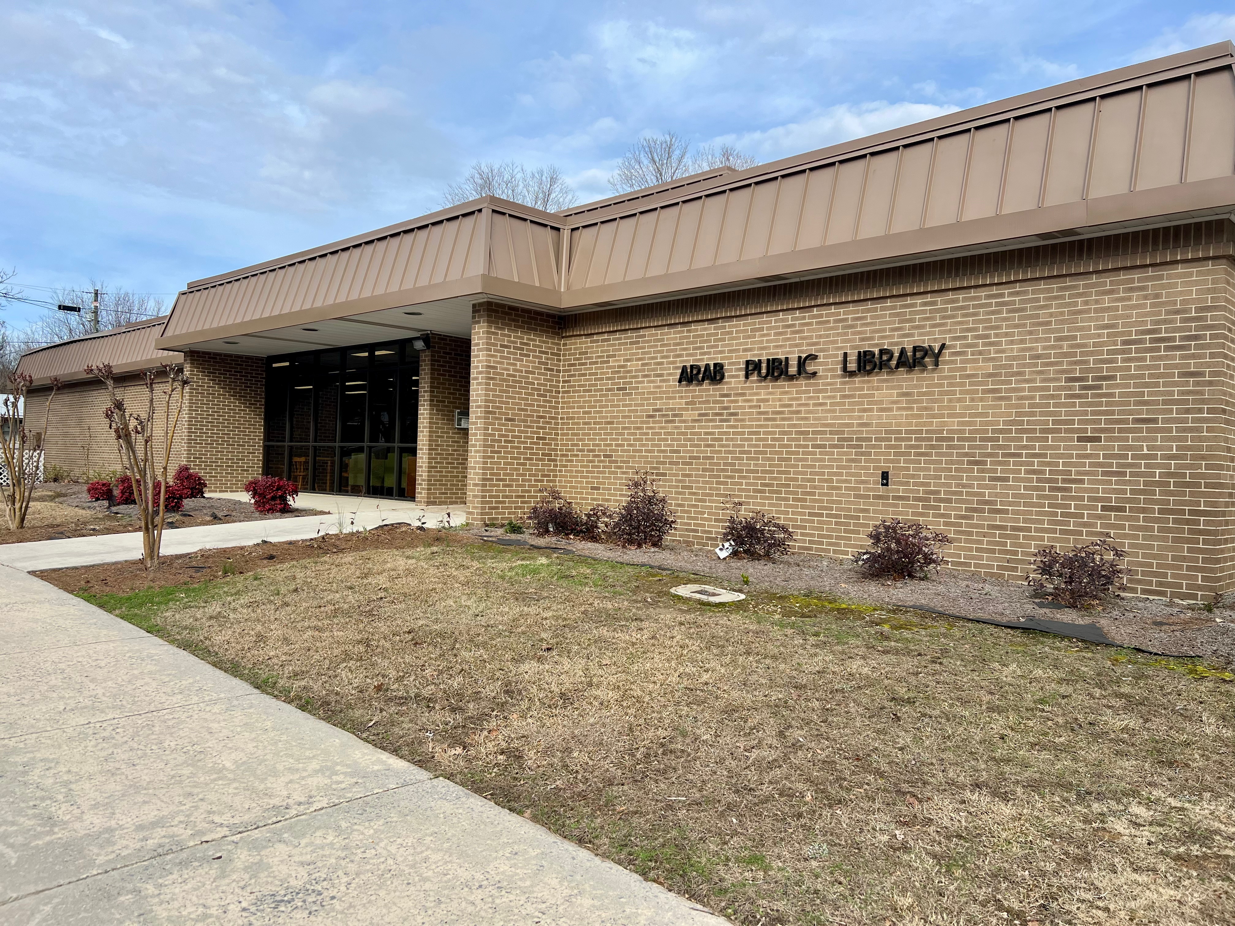 An exterior shot of the Arab Public Library shows a tan brick building with a tan metal roof and a cloudy sky above it.